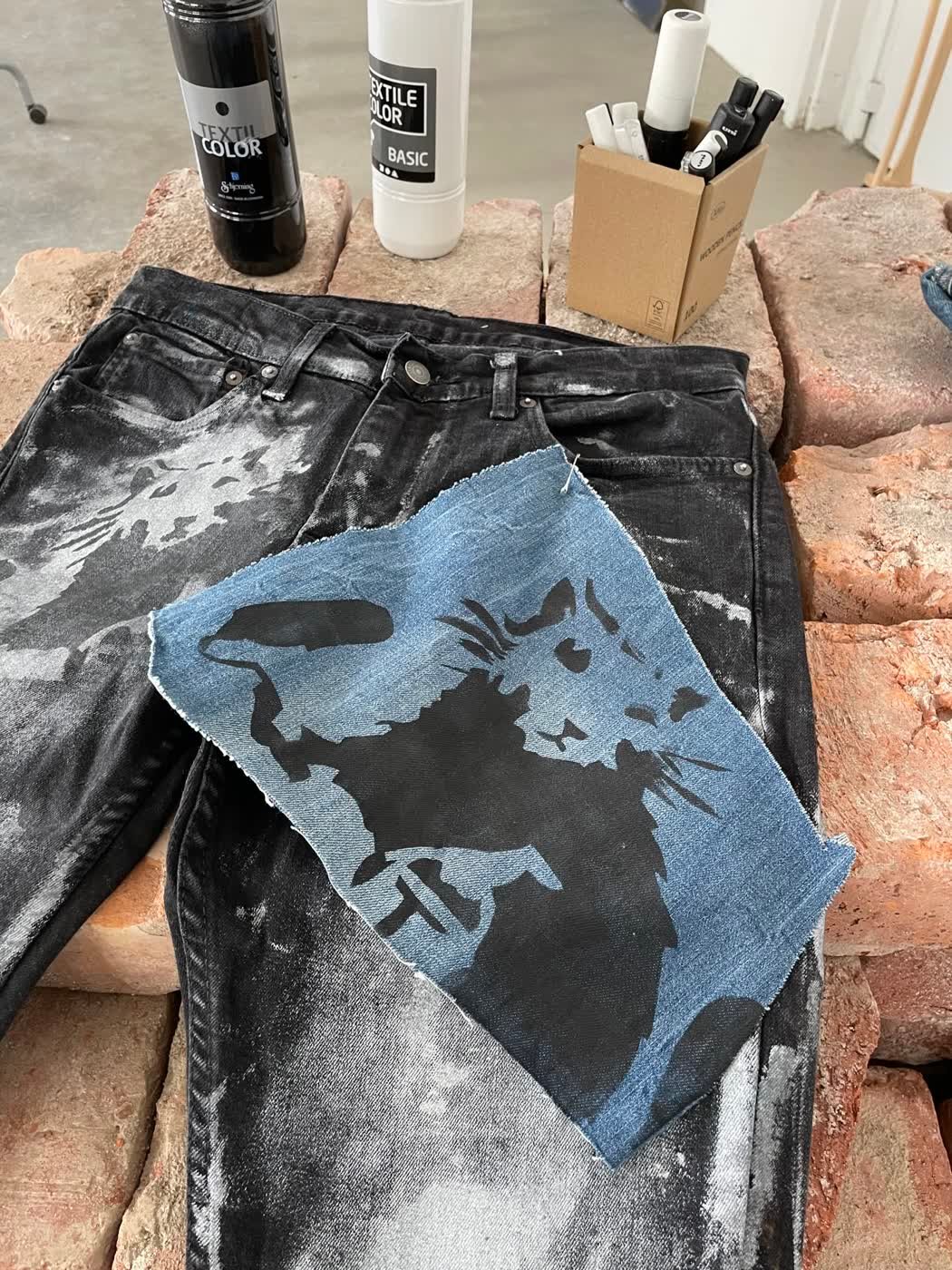 Bleach-painted black jeans with a raw-edge blue denim patch, laid over reclaimed bricks.