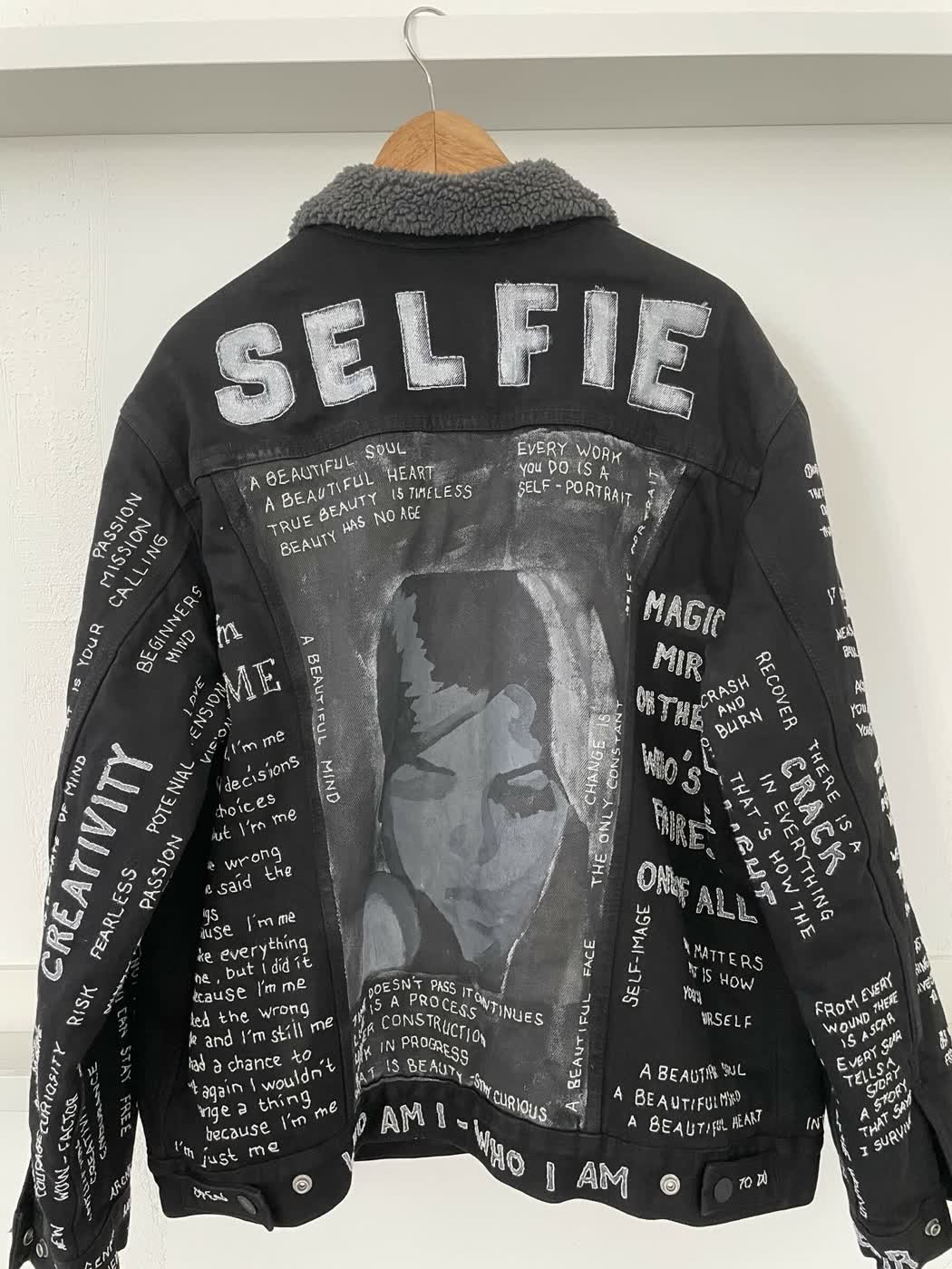 Black denim jacket with a painted grayscale portrait and white slogans including "Selfie" and "A beautiful mind".