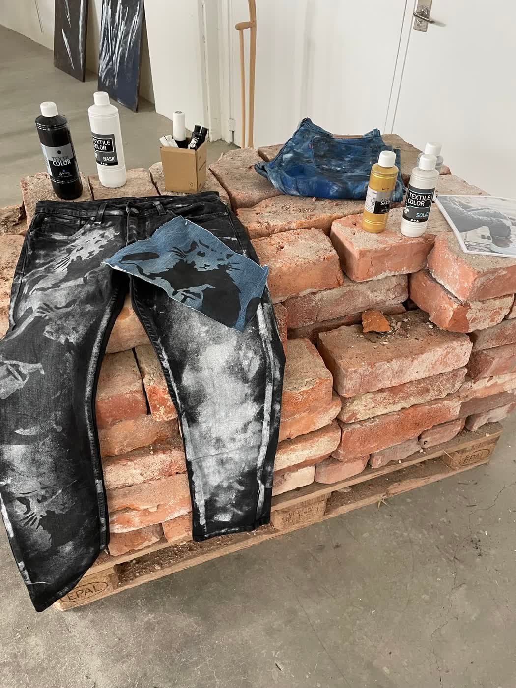 Customised black-and-silver bleached jeans laid over a stack of reclaimed bricks beside paint bottles.