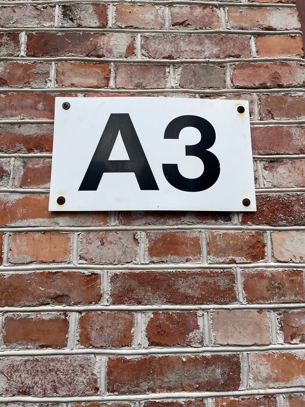 A white enamel "A3" plate bolted to the workshop's red-brick wall - the unit marker to look for.