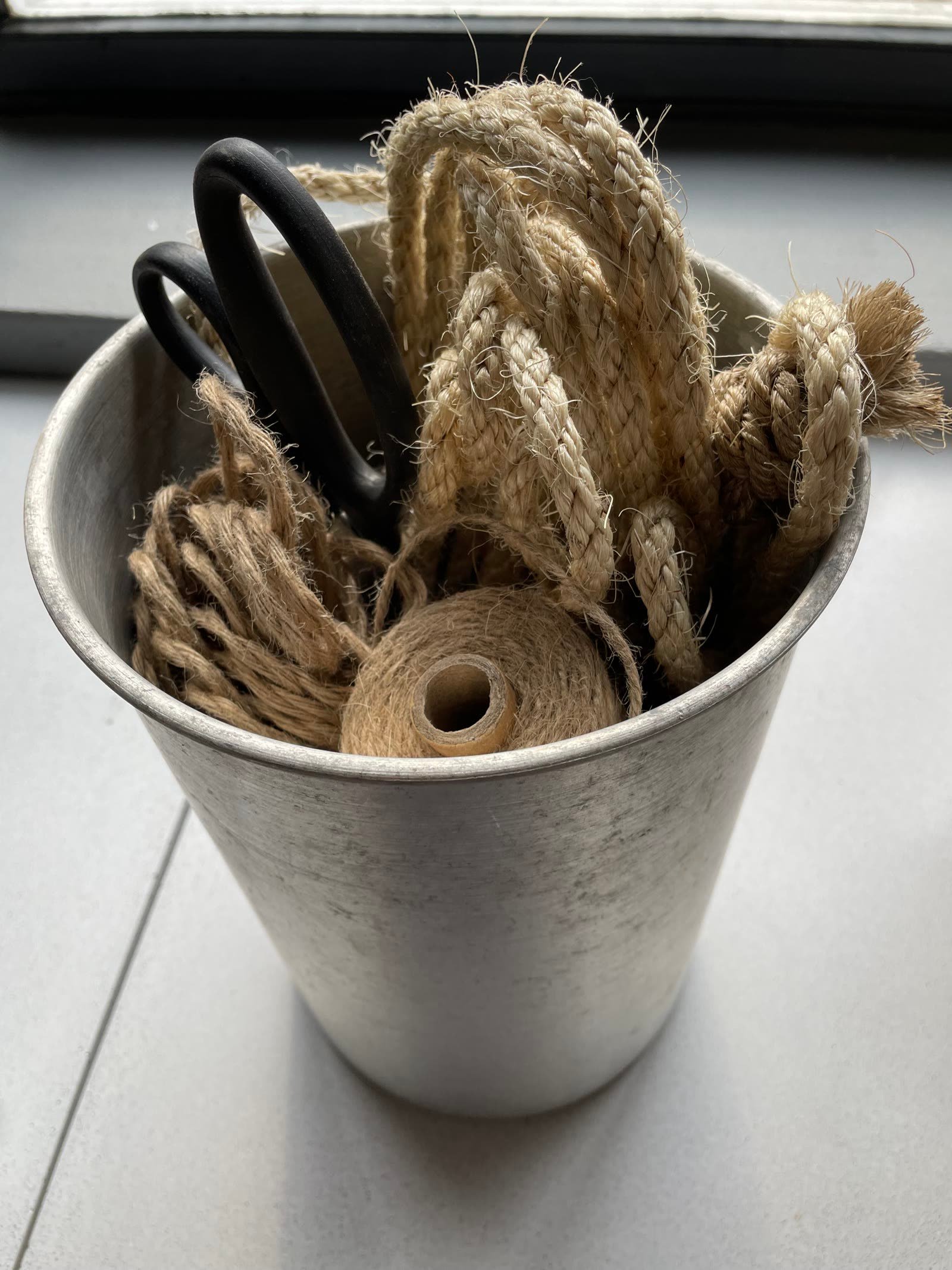 A metal pail of jute rope, cord and a large pair of workshop scissors on the windowsill.
